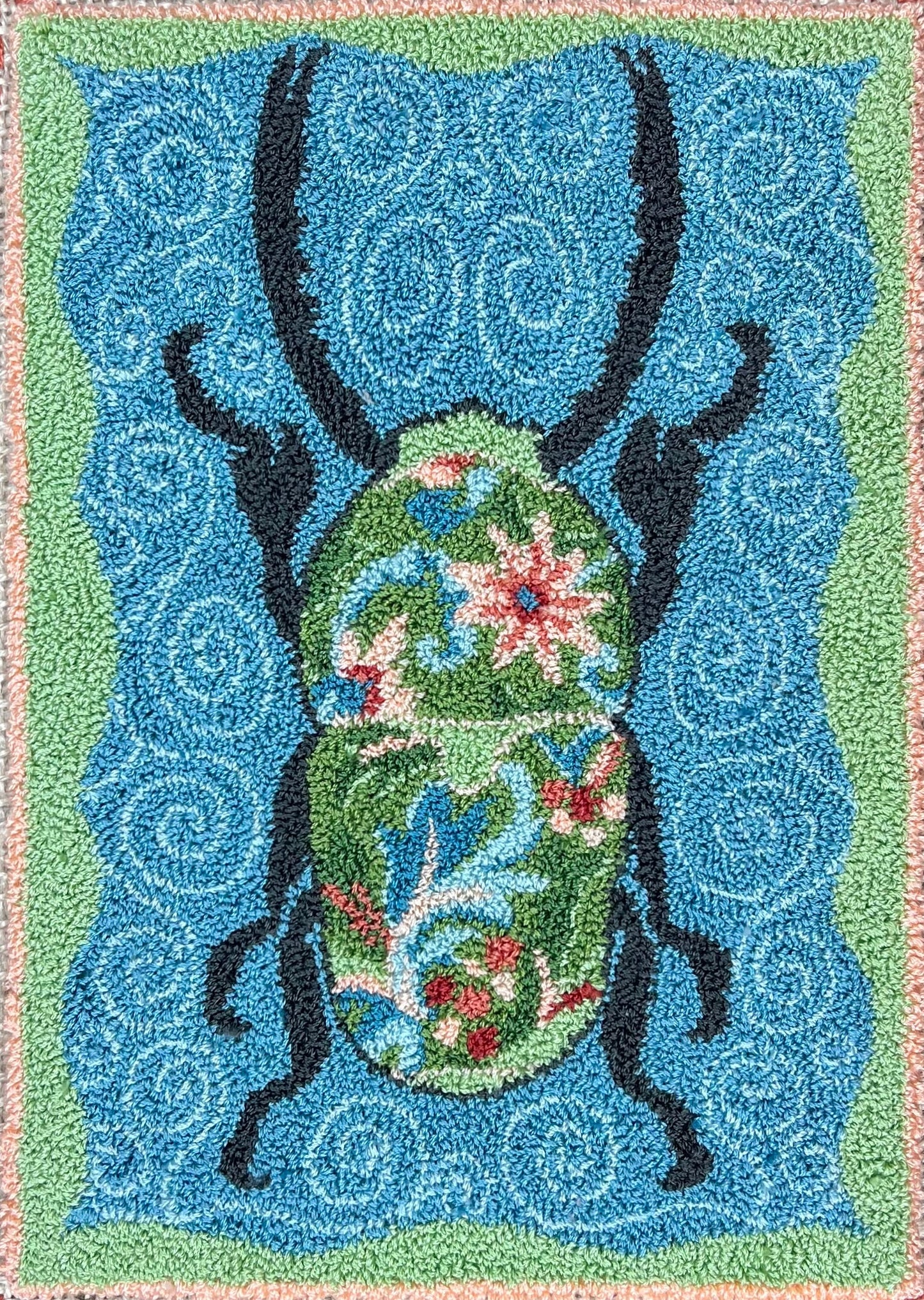 A paper rug hooking pattern featuring a stylized floral design with a prominent black beetle on a blue background, surrounded by a multicolored border.