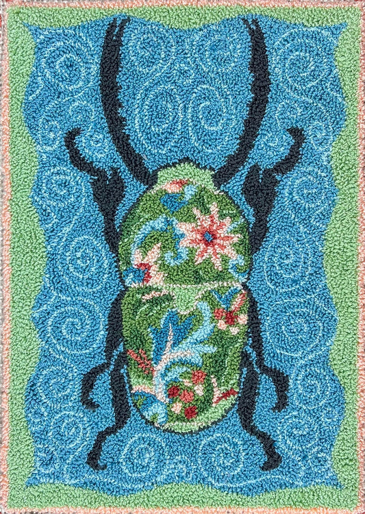 A paper rug hooking pattern featuring a stylized floral design with a prominent black beetle on a blue background, surrounded by a multicolored border.